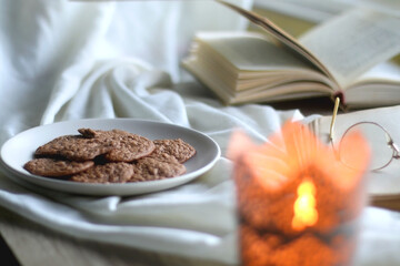 Lit candle, open books, reading glasses and plate of gluten free cookies on the table. Hygge at home. Selective focus.