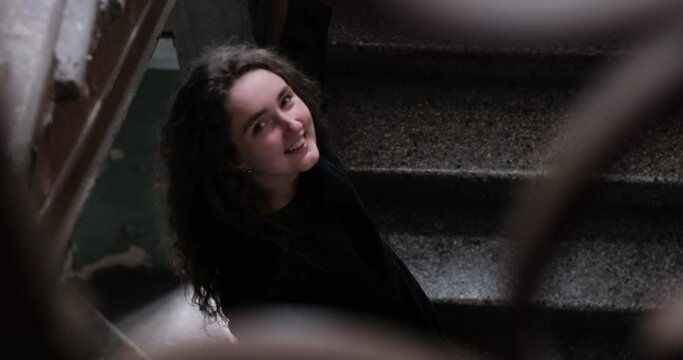 Cute girl with long curly hair on the stairs indoors. Shy gentle girl looks into the camera