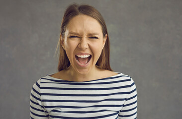 Obraz premium Close up studio shot of a crazy stressed beautiful young woman with an angry displeased face expression screaming loudly at the camera. Portrait of a mad girlfriend or enraged school teacher
