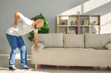 Young man leaning on sofa at home is holding his lower back due to severe pain and cramps. Concept...