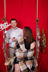 Obraz premium happy couple hugging on wooden swing decorated with christmas tree branches on red background. cozy young family.