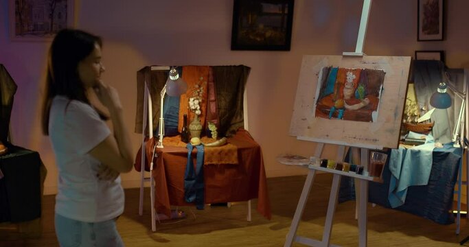 Young woman artist is pensively looking at an easel with a painting. Woman admires a still life on canvas. Artist's studio with easels, canvases and paints, dim lights. 4k, ProRes
