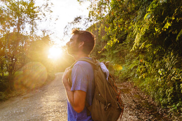Horizontal portrait of young latin american man hiking at sunset. Horizontal view of traveler man with backpack walking in highway with lens flare. People and travel destination in South America.