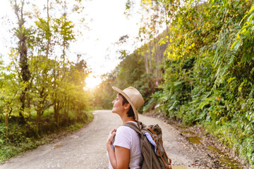 Cheerful caucasian woman with short hair and country hat hiking with forest background. Horizontal view of carefree woman in the highway with backpack traveling in nature. People and travel concept.