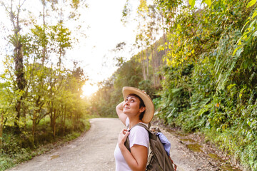 Cheerful caucasian woman with country hat looking up and hiking with forest background. Horizontal view of carefree woman in the highway with backpack traveling in nature. People and travel concept.
