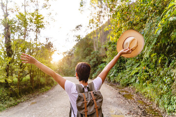 Cheerful caucasian woman with arms wide open holding hat hiking with forest background. Horizontal view of carefree woman in the highway with backpack traveling in nature. People and travel concept.