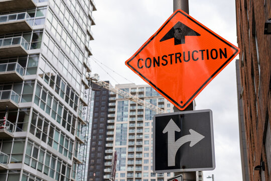 Construction orange sign near construction site against tall buildings and sky