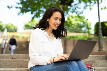 Young businesswoman sitting on steps outdoors and working on laptop. Beautiful girl learning in the park