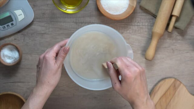Woman prepairing dough mixing the ingredients (water, sugar, yeast) in a bowl using a wooden spoon. Step 1 of process of kneading yeast dough. Top view, flat lay.