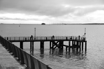 black and white image of old wooden pier. People fishing on pier over large river.