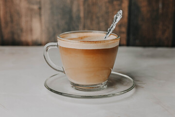 Coffee with milk in a transparent cup with an embossed spoon on a plate on a dark wooden background