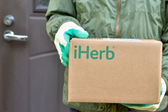 Logo iHerb on cardboard box in courier hands. iHerb is an herbal products company, provides and delivers health and wellness products to people around the world: Riga, Latvia - September 10, 2021