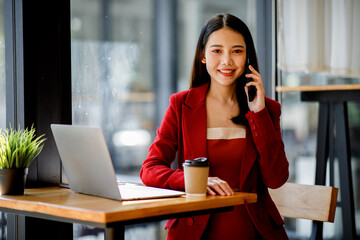 Asian Young businesswoman wearing red suit using a laptop computer. Female working on laptop in an in the cafe.