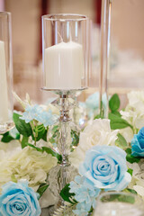 Candles in transparent glass jars on a wooden table A glass vase with a candle inside decorated with white and blue roses and greenery on the table Wedding table set decor Close up