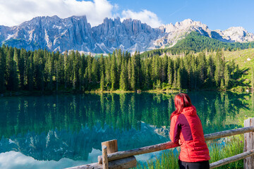 Woman enjoying the turquoise Lago di Carezza 1,925m altitude (mountain lake) view as he has mountain walk in Dolomite Mountains, Italy. Active people in nature concept. - obrazy, fototapety, plakaty