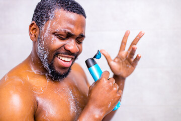 Attractive young cheerful man singing while washing in the shower, holding shampoo bottle like...