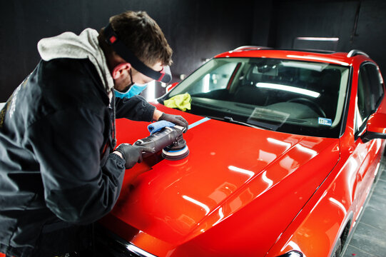 Car Detailing Concept. Man In Face Mask With Orbital Polisher In Repair Shop Polishing Orange Suv Car.