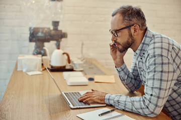Bearded man talking on cellphone and using laptop in cafe