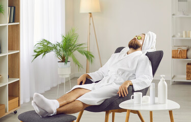 Calm man in bathrobe and towel after shower relax in chair do beauty facial procedures. Metrosexual guy make face skincare mask treatment for glowing skin, have spa day on weekend at home. - obrazy, fototapety, plakaty