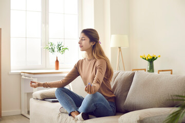 Fototapeta Calm millennial girl sit on sofa with mudra hands meditate practice yoga at home. Relaxed young woman do physical activity relieve negative emotions breathe fresh air. Meditation concept. obraz