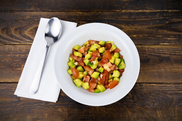 White plate with salad of avocado and tomato, spoon on white napkin on brown wooden table, top view, flatlay