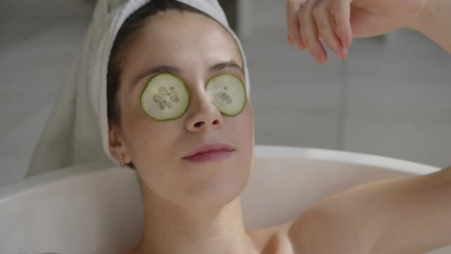 Closeup shot of relaxed bautiful caucasian young woman lying in spa bath and putting cucumber on her eyes with white towel on head, eyes closed. Relaxing spa wellness treatments at home