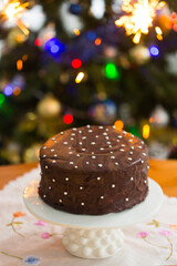 Homemade holiday chocolate cake with tiny sugar stars on white cake stand on the background of a Christmas tree with glowing garlands