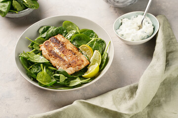 A plate with fresh green raw spinach and fried wild salmon, lime slices, cream cheese sauce on grey concrete surface