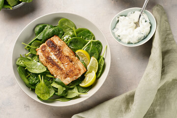 A plate with fresh green raw spinach and fried wild salmon, lime slices, cream cheese sauce on grey concrete surface