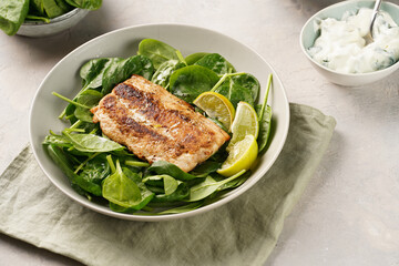 A plate with fresh green raw spinach and fried wild salmon, lime slices, cream cheese sauce on grey concrete surface