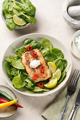 A plate with fresh green raw spinach and fried wild salmon, lime slices, cream cheese sauce, red chilli pepper on grey concrete surface