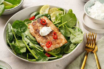 A plate with fresh green raw spinach and fried wild salmon, lime slices, cream cheese sauce, red chilli pepper on grey concrete surface