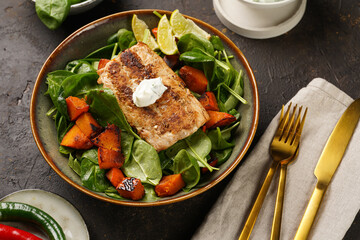 A plate with fresh green raw spinach and fried wild salmon, pumkin pieces, lime slices, cream cheese sauce, red chilli pepper on brown surface