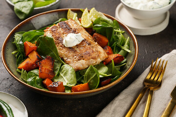 A plate with fresh green raw spinach and fried wild salmon, pumkin pieces, lime slices, cream cheese sauce, red chilli pepper on brown surface