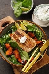A plate with fresh green raw spinach and fried wild salmon, pumkin pieces, lime slices, cream cheese sauce, red chilli pepper on brown surface