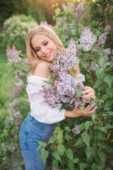 Obraz na płótnie Canvas a woman with blond hair in a white blouse and jeans hugs a lilac bush