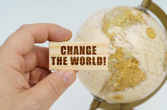 A man holds in his hand a wooden plate on which it is written - Change The World. In the background is a globe.