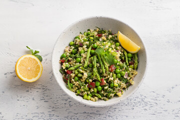 Traditional middle eastern or arabic tabbouleh salad of bulgur, tomatoes, asparagus, green peas and greens with half lemon on light gray background, top view. Healthy homemade vegan food
