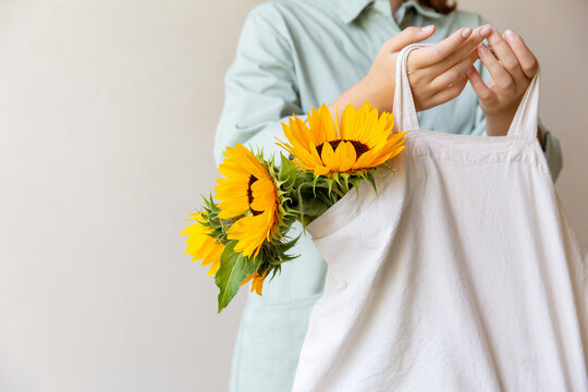 Eco-natural cotton reusable bag with sunflowers, hanging on the arm of a girl in a light green jumpsuit, on a light background. The concept of an ecological lifestyle and clothing. copy space
