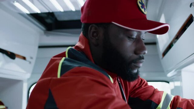 Tracking shot of African American doctor in red uniform using defibrillator on patient with heart attack while working in emergency service vehicle