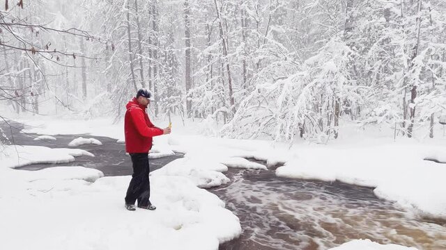 The photographer does to a photo and video the wild nature at snow storm by smartphone, he is dressed in red color jacket, the wild frozen small river in the winter wood, ice, snow-covered trees