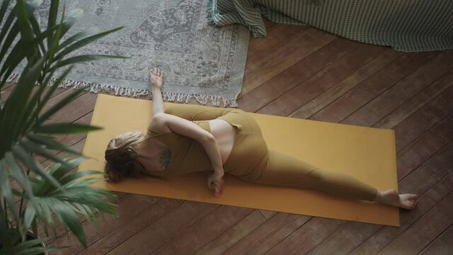 Slim fair haired young woman in yellow sporty top and leggings does yoga poses on mat lying on parquet floor near bed and in bedroom top view