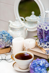 A visual for content. Still life in vintage style. A mug with a drink, an old book, a cage, candles and hydrangea flowers in the garden on a white wooden table. The concept of a tea ceremony.