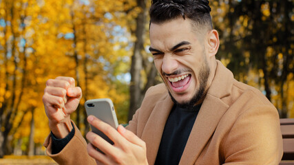 Excited hispanic guy reading good news on smartphone sitting on bench in autumn park young man...
