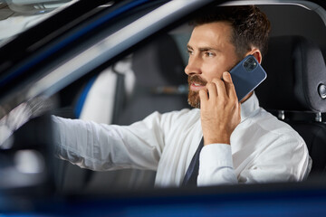 Successful businessman talking on mobile while examining modern car at auto salon. Male customer in suit buying new vehicle. Technology and transport concept. 