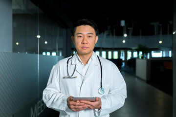 Concentrated and confident, Asian doctor, in the corridor of the clinic, looks at the camera and holds a tablet computer