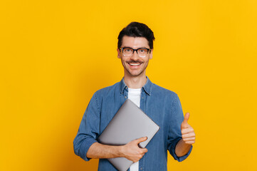 Attractive smart caucasian guy, with glasses, student or freelancer, holding a laptop in hand, standing against isolated orange background, looking at camera, smiling friendly, shows thumb up gesture