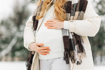 pregnant woman in warm winter clothes standing outside on a snowy winter day, with hands on her...