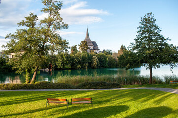 Naklejka premium Sunlit church in the village of Maria Worth on the shores of Lake Worthersee in the Austrian mountains. Irradiated grass in the park and two benches. Beautiful lake with clear blue water.