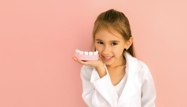 Cute little baby girl demonstrates a healthy snow-white smile, holding a model of a jaw in her hand to demonstrate proper brushing of teeth. International Dentist's Day. A child in a medical gown - Powered by Adobe